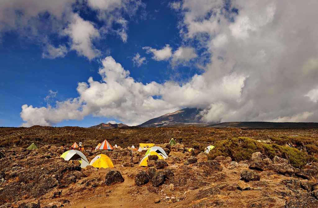 A scatter of small yellow, white and green dome tents pitched among red-brown rocks and low scrub, with the cloud-wrapped summit cone rising beyond under a deep blue sky.