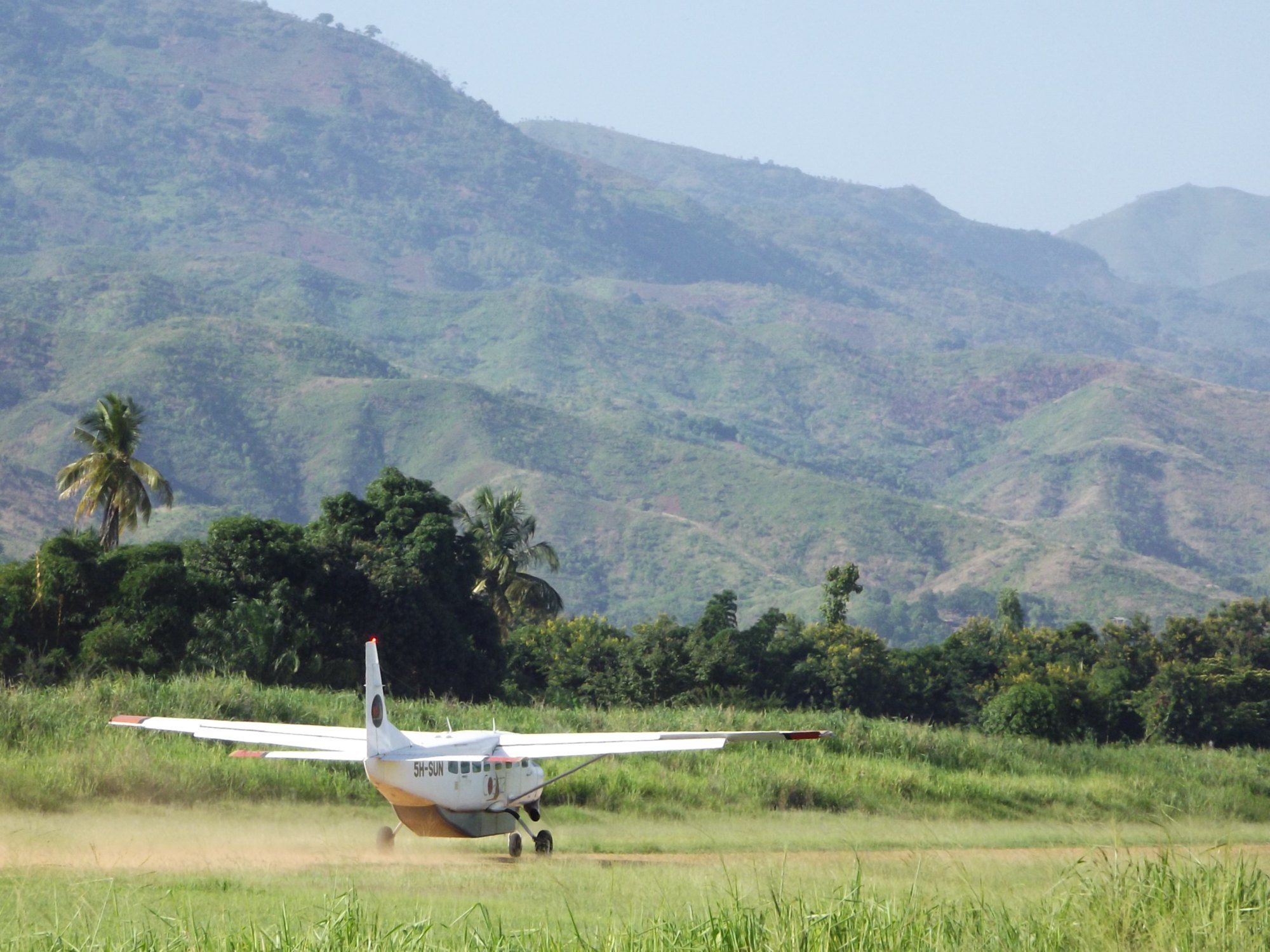 A single-engined light aircraft standing on a mown grass airstrip cut out of green bush, with a palm and dense trees along the edge and a wall of steep forested mountains rising into haze behind it.