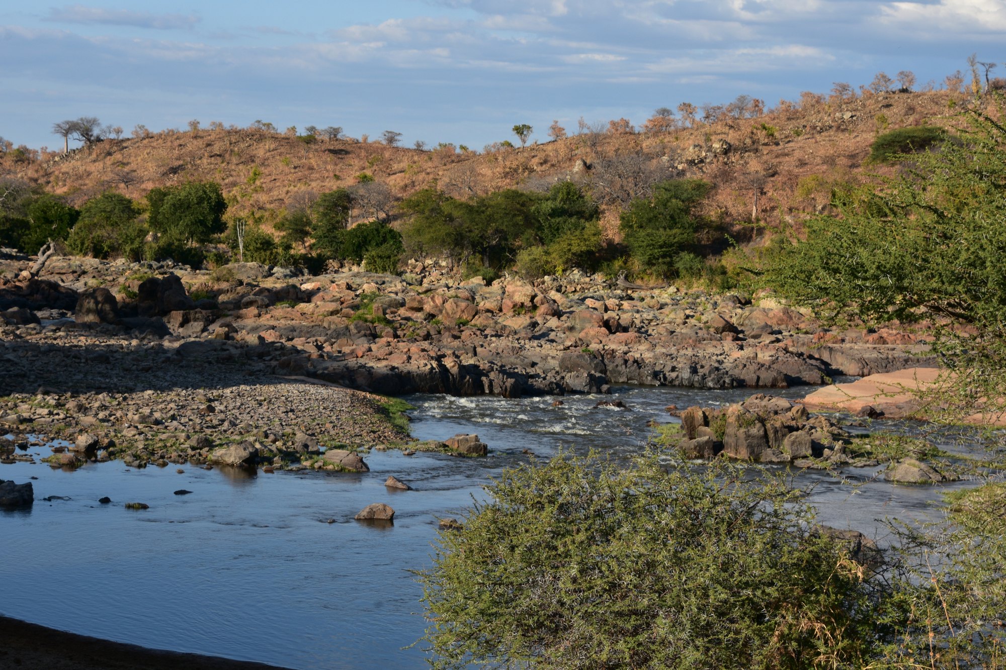 The Great Ruaha River running shallow over a bed of dark boulders, with dry bush and a low rocky ridge rising behind it under a blue sky.