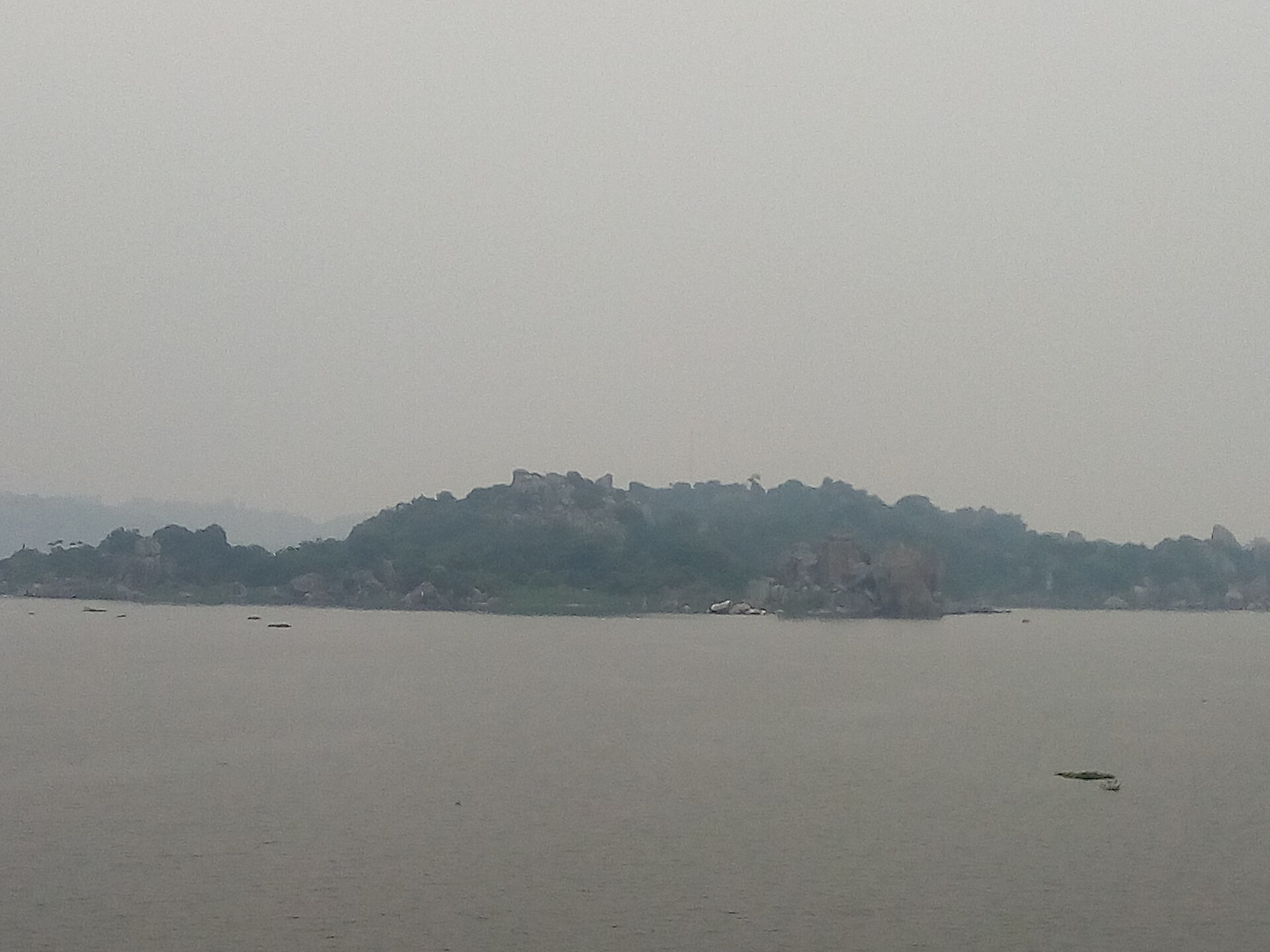 The low rocky island seen across flat grey water in soft haze, its granite boulders and scrub showing as a dark silhouette.