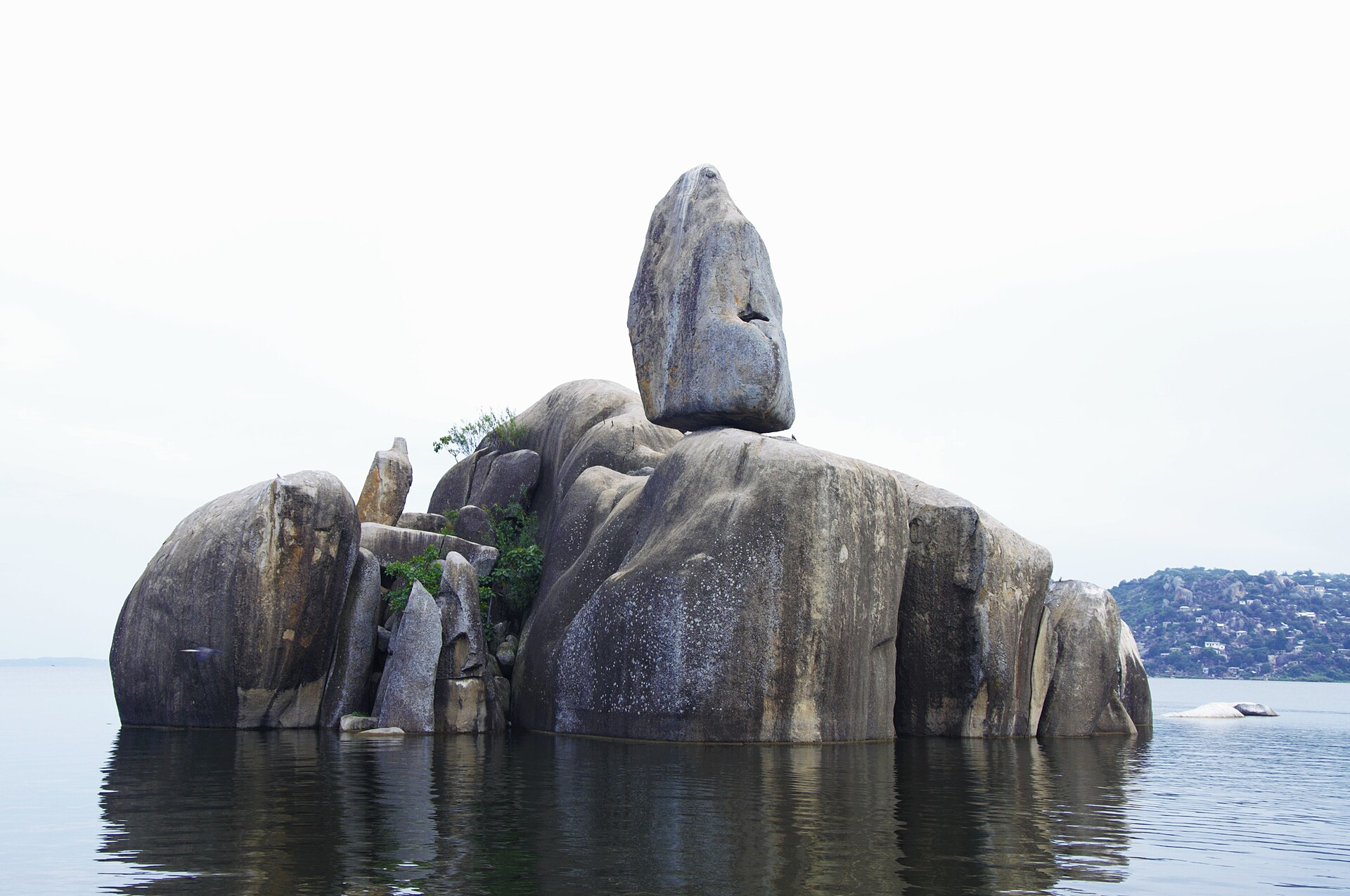 A tall balancing boulder perched on a stack of granite rocks in the shallows of Lake Victoria, with a few small trees growing between them.