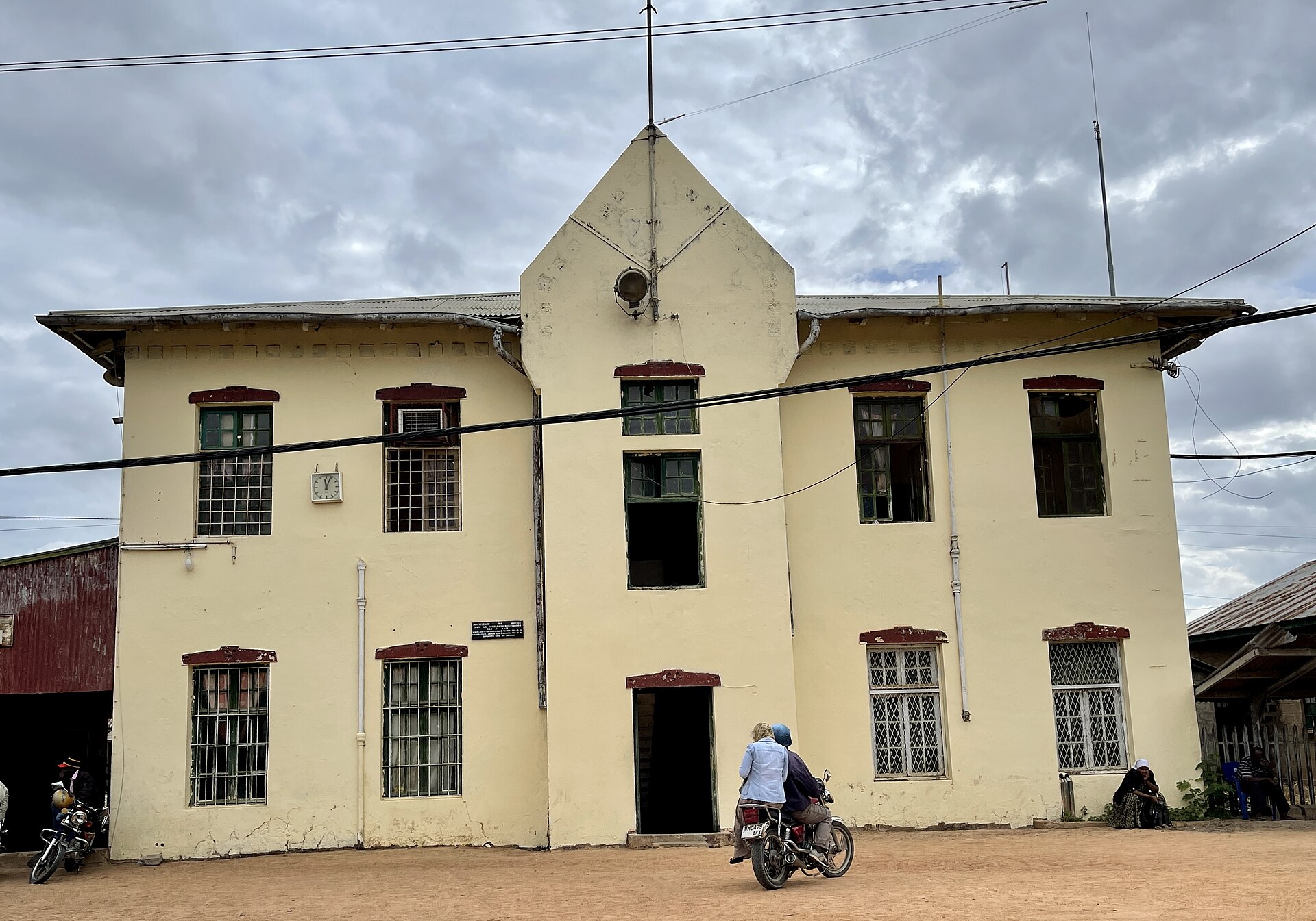The cream-painted colonial railway station with its steep gable and shuttered windows, a motorcycle and two people passing the front door.