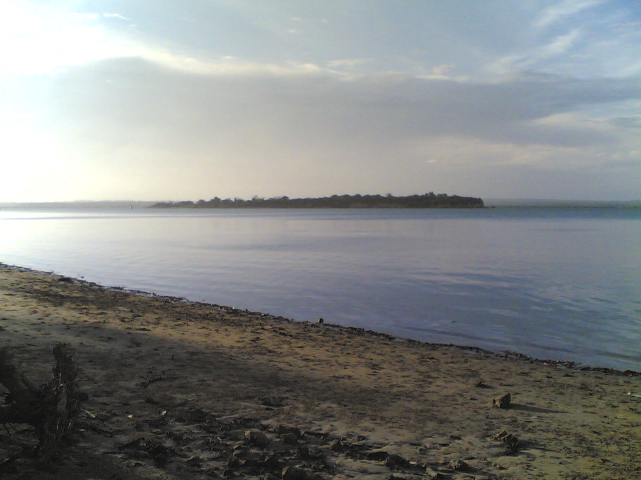 Toten Island lying low and wooded on the far side of flat calm water, seen from a sand and shingle shore in Tanga bay under a pale hazy sky.