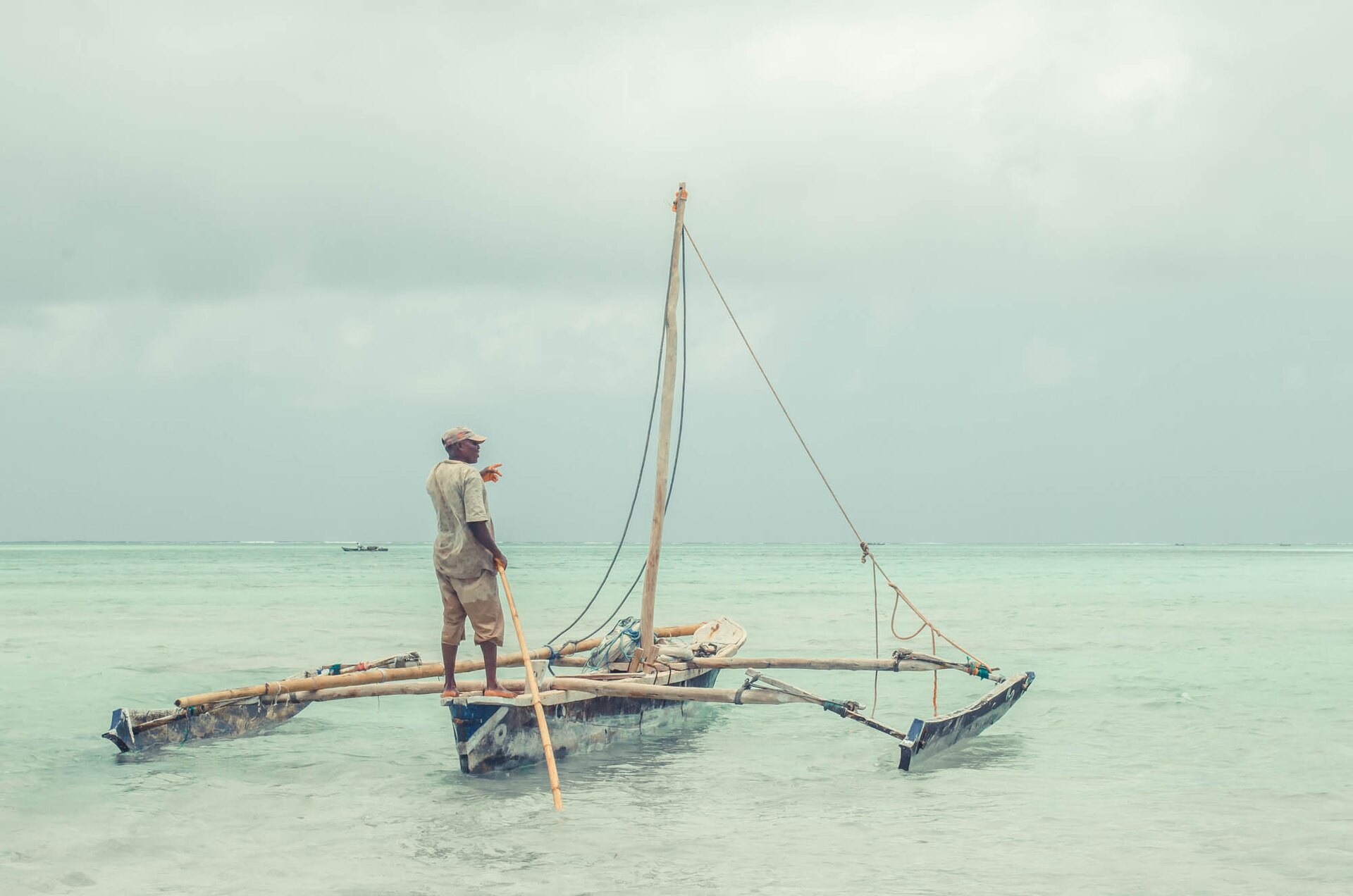 A traditional outrigger sailing dhow on flat, milky-green water off Makunduchi, a single figure standing at the mast against a pale hazy sky.