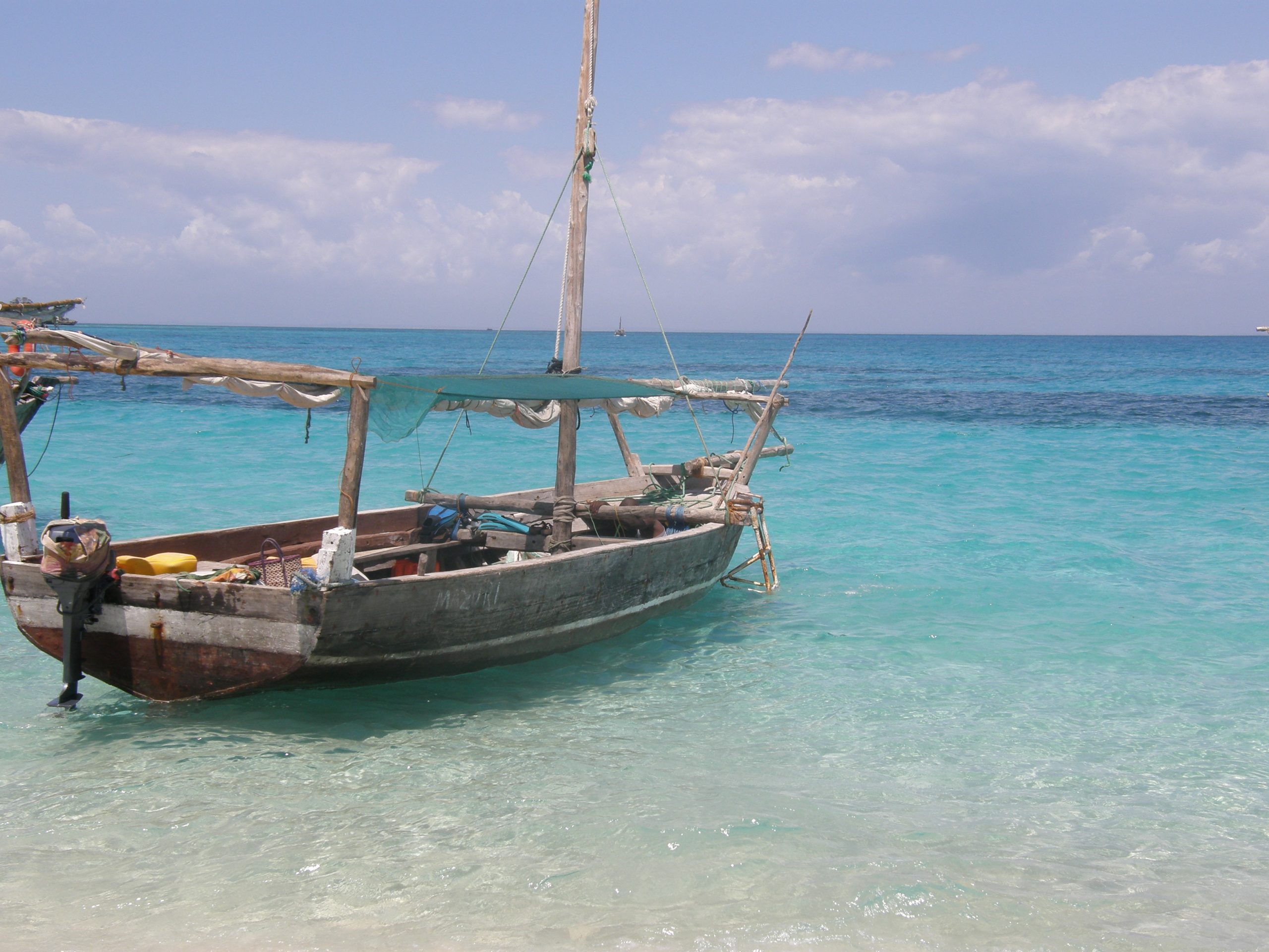 A wooden dhow with a canvas awning and snorkelling fins aboard, moored in clear turquoise shallows in Menai Bay off south-west Unguja, with the darker water of the reef edge behind it.